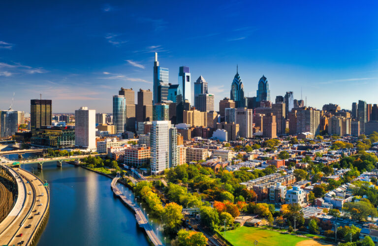 Philadelphia skyline aerial view with the Schuylkill River, Schuylkill Expressway and residential neighborhoods and autumn colored trees in the foreground, and a blue sky with clouds in the background.