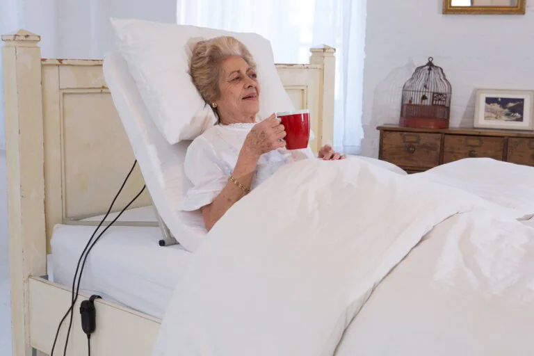`Elderly woman drinking from red mug while resting in adjustable bed`