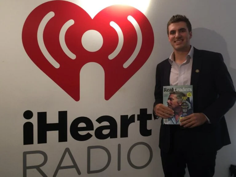 Man holding a “Real Leaders” magazine in front of the iHeartRadio logo wall.