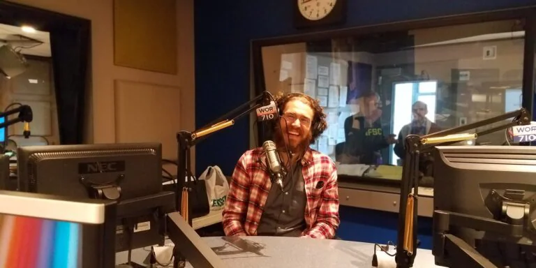 Smiling man in a plaid shirt wearing headphones at a WOR 710 radio studio microphone.