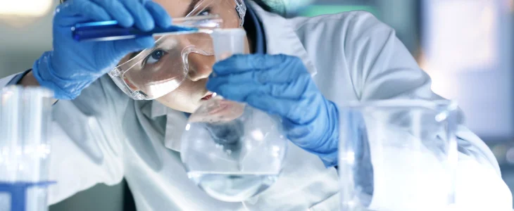 A scientist in a lab coat and goggles pours a blue liquid into a flask.
